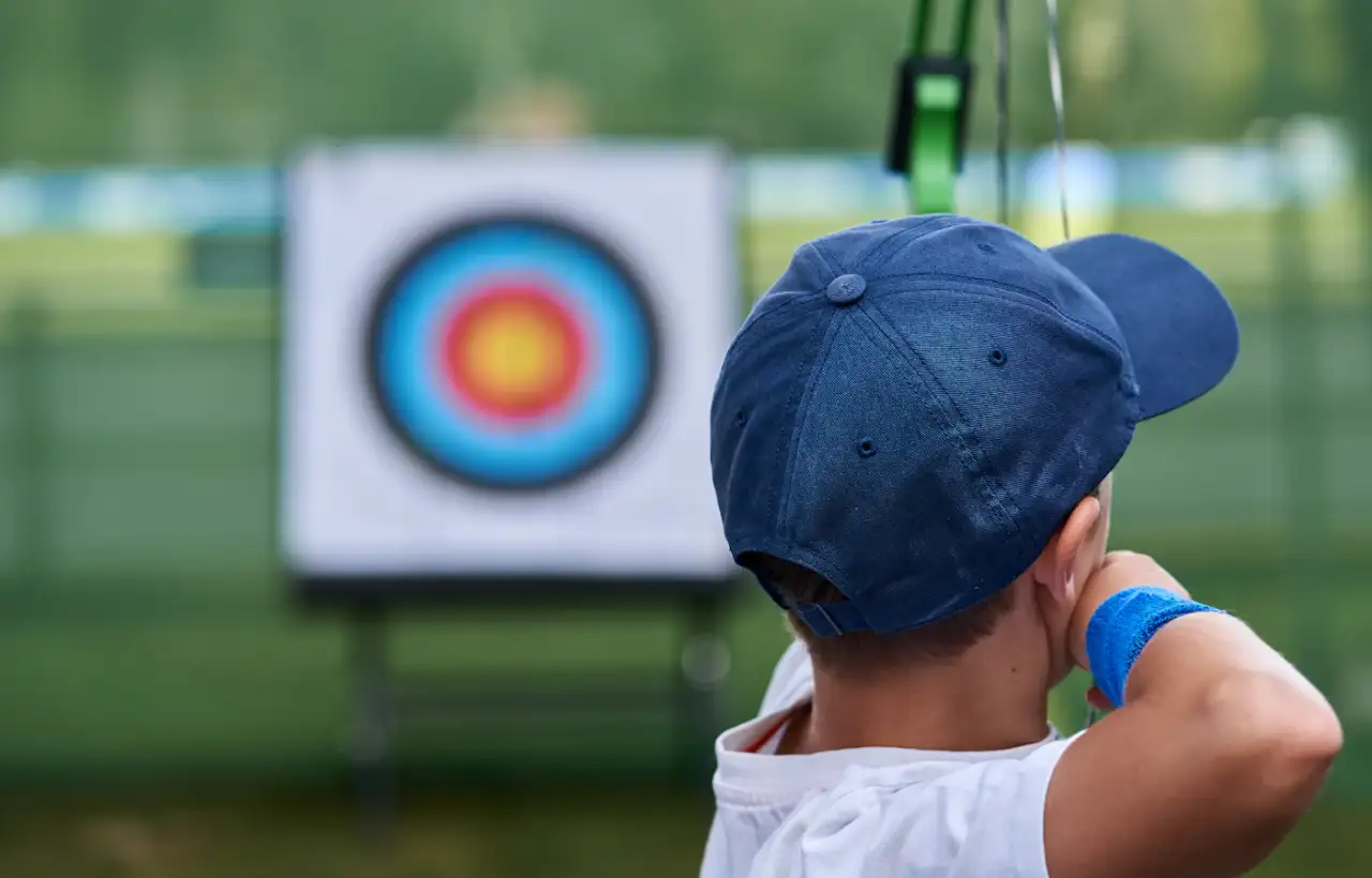A young Scout in a cap drawing a bow and aiming at an archery target