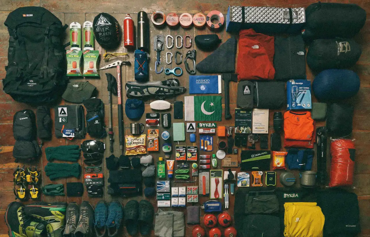 Camping gear including a backpack, water bottle, flashlight and clothing laid out on the ground